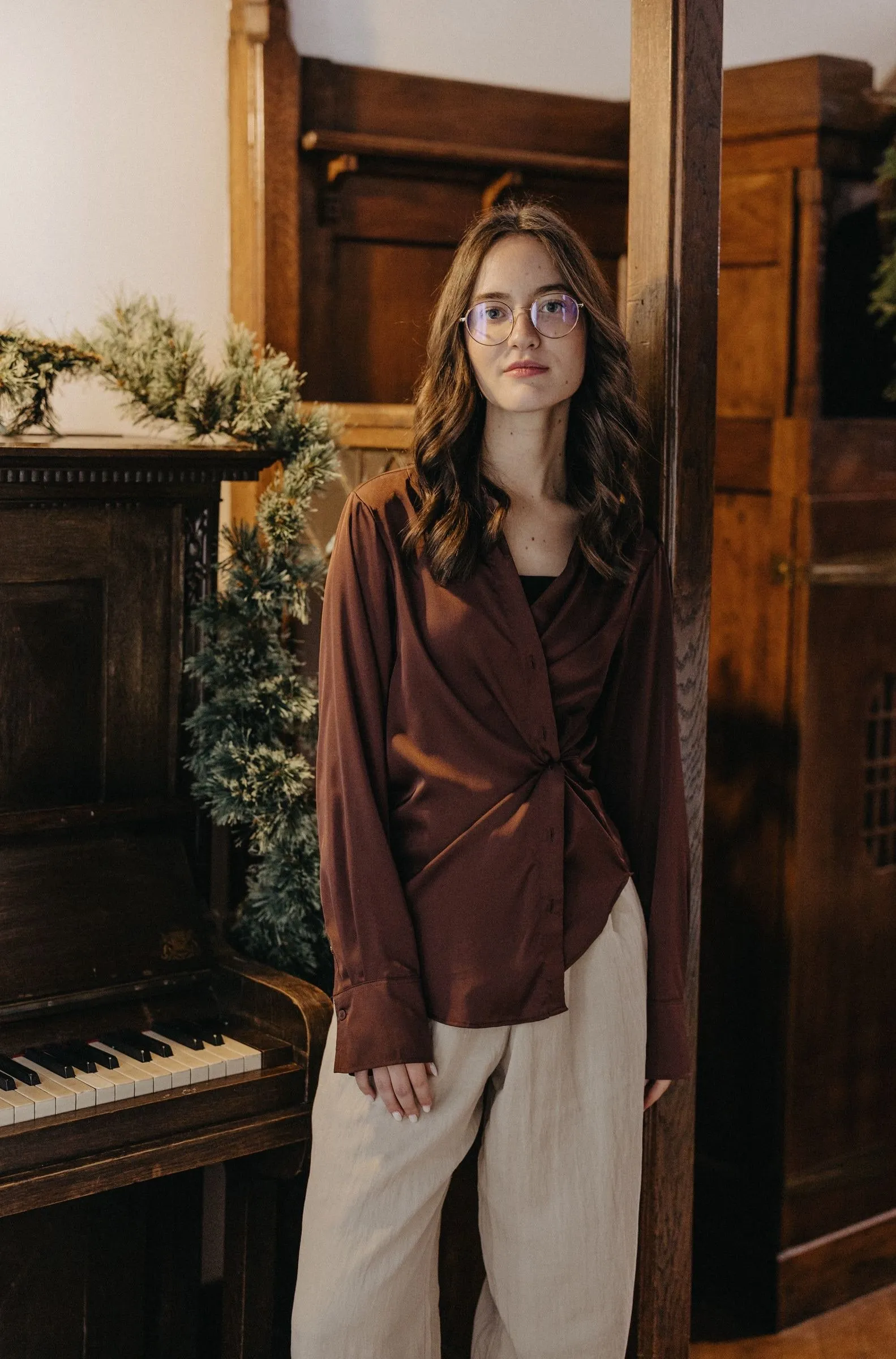 Sara Toadere, photographer, in glasses and a brown satin blouse, leaning against a wooden post beside an old upright piano decorated with a pine garland