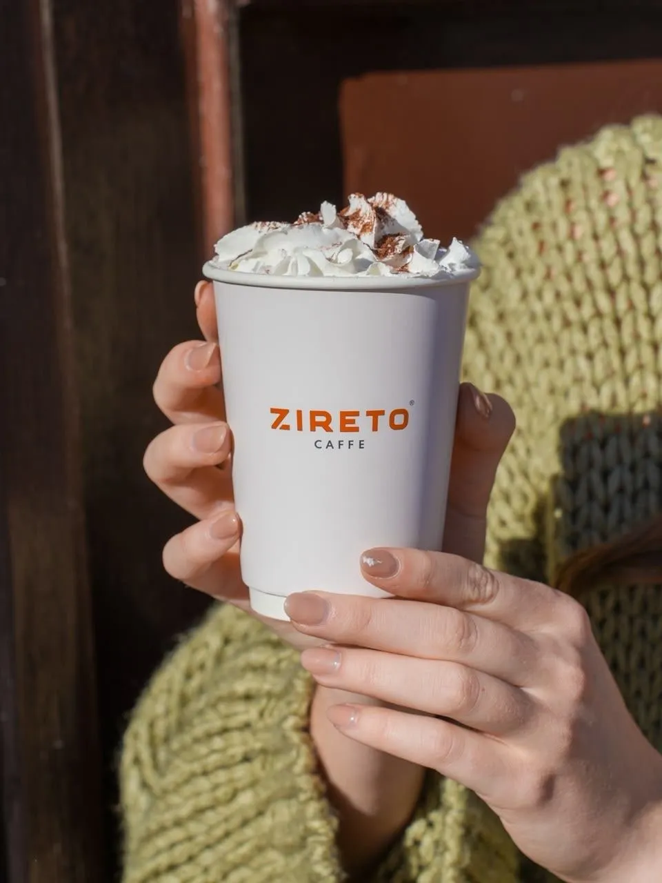 Două mâini țin un pahar alb de cafea ZIRETO caffe cu frișcă și cacao, în fața unei uși de lemn închise la culoare, cu mâneca unui pulover verde tricotat în spate