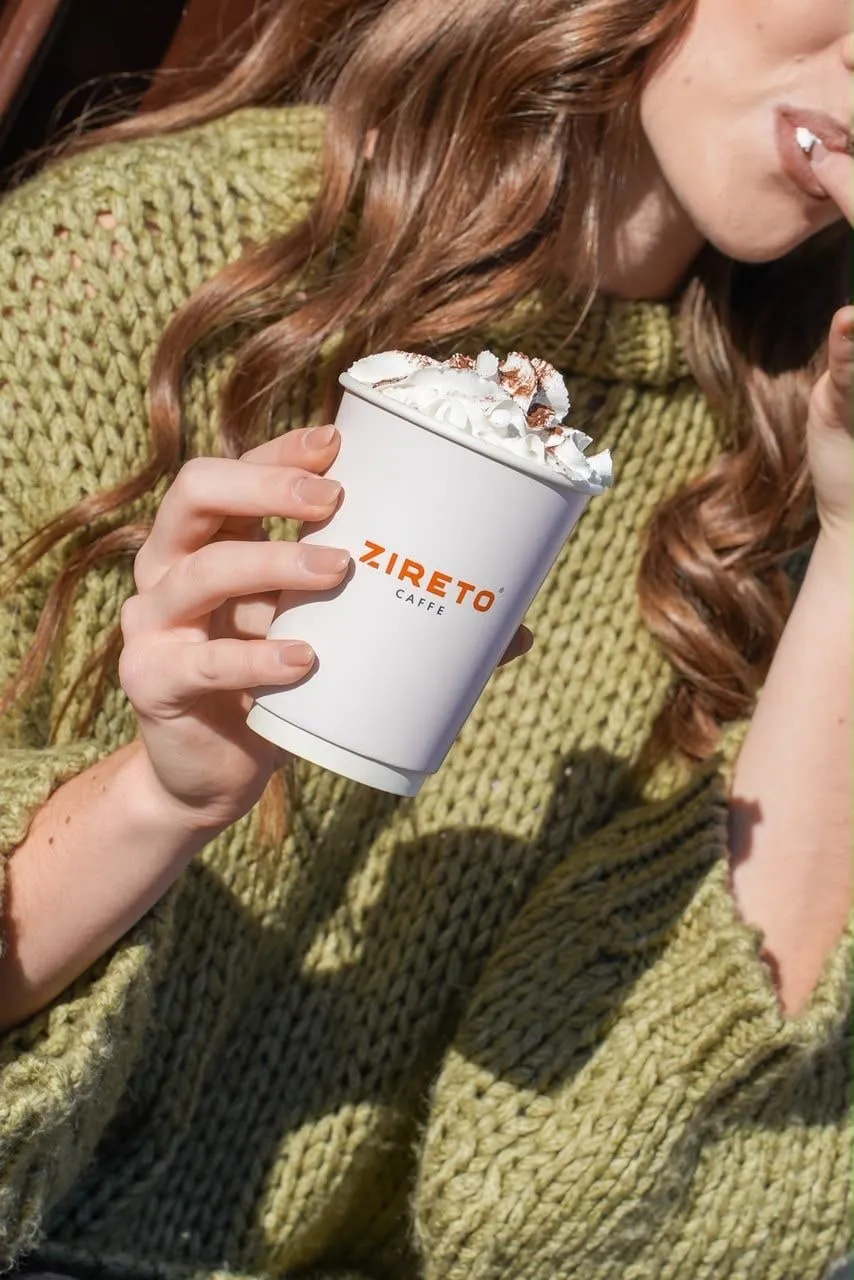 Un pahar alb de cafea ZIRETO caffe cu frișcă, ținut în fața unui pulover verde tricotat, cu părul lung, ondulat și buzele în cadru