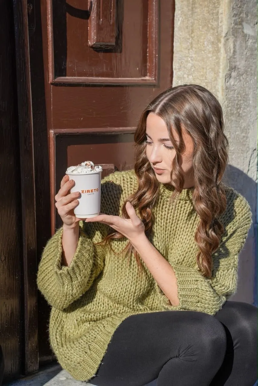 O tânără în pulover verde tricotat, așezată lângă o ușă maro de lemn, privește paharul de cafea cu frișcă din mâna ei