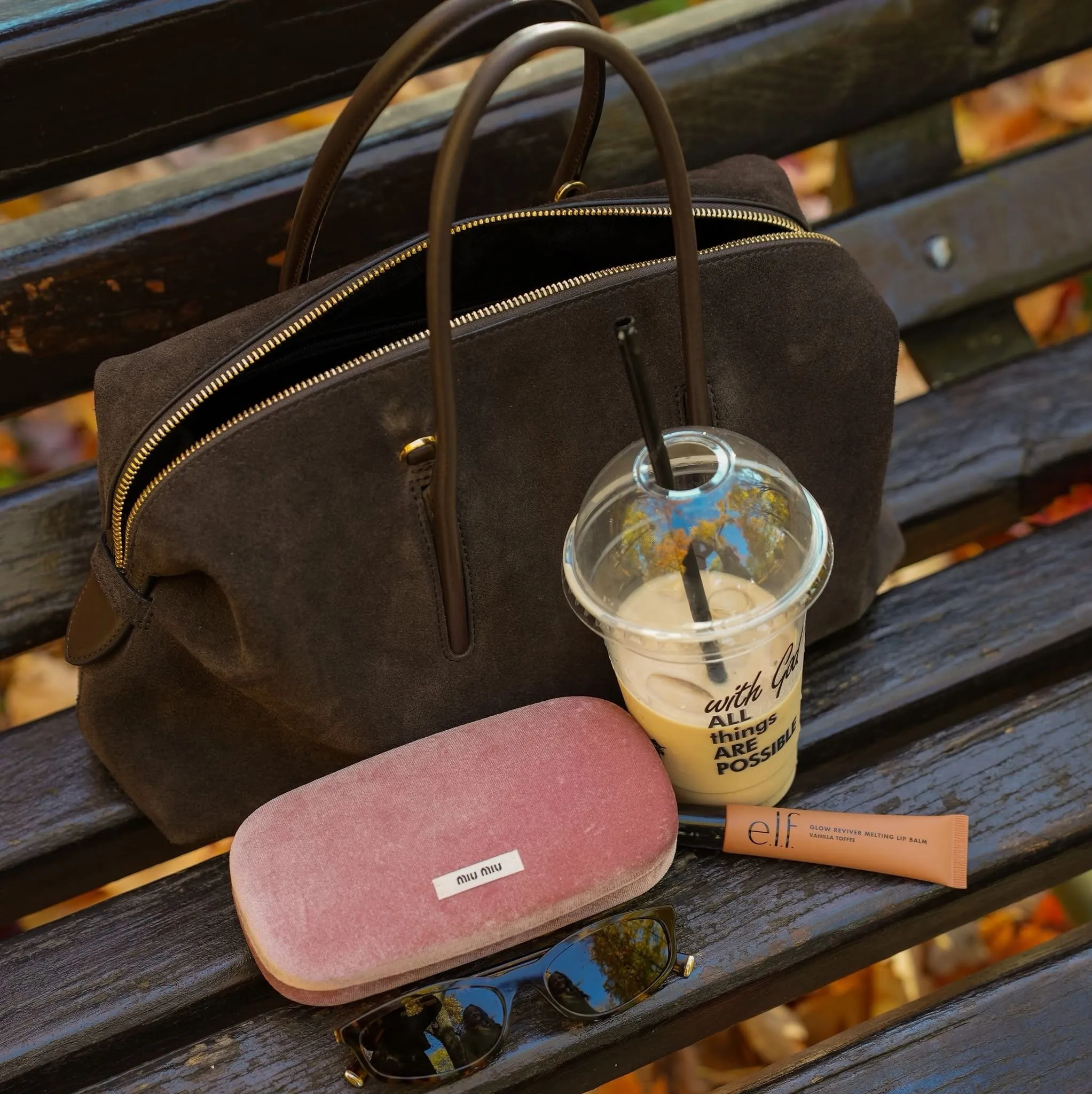 Detaliu pe o bancă: o geantă din piele întoarsă, o cafea cu gheață, o cutie roz de ochelari Miu Miu, un balsam de buze e.l.f. și ochelari de soare
