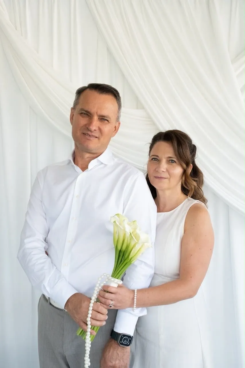 Un bărbat și o femeie îmbrăcați în alb, unul lângă altul în fața unei draperii albe, țin împreună un buchet de cale albe cu un șirag de perle