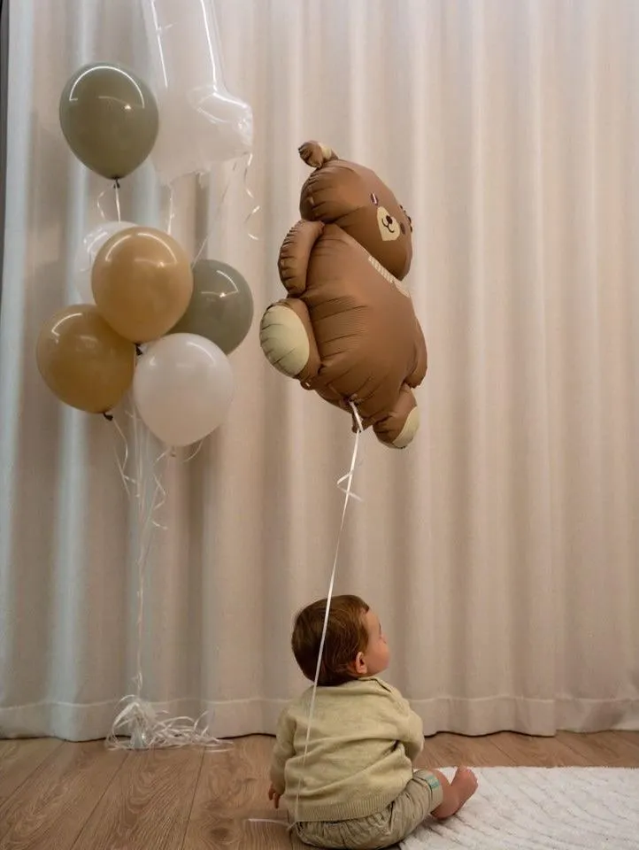 Un bebeluș așezat pe parchet, văzut din spate, privind în sus spre un balon în formă de ursuleț, lângă un buchet de baloane verzi-gri, bej și albe, în fața unei draperii deschise la culoare