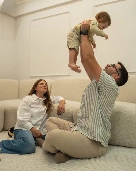 A man sitting on a rug lifts a baby high above his head while a woman sitting on the floor beside him looks up smiling, in front of a cream sofa
