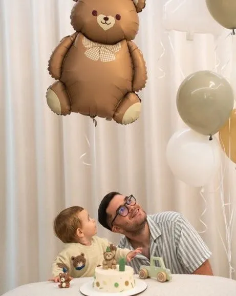 A man and a toddler at a table with a small cake topped with a green “1” candle and a teddy-bear figure, under a large teddy-bear balloon