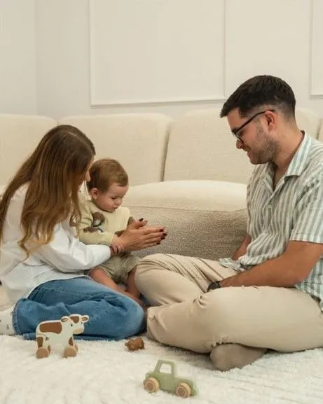 A woman holding a toddler and a man sitting on a white rug in front of a cream sofa, with wooden toys on the rug: a cow and a little car