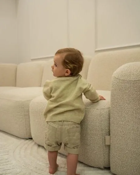 A toddler in a pale green jumper standing at a cream sofa, seen from behind, head turned to one side