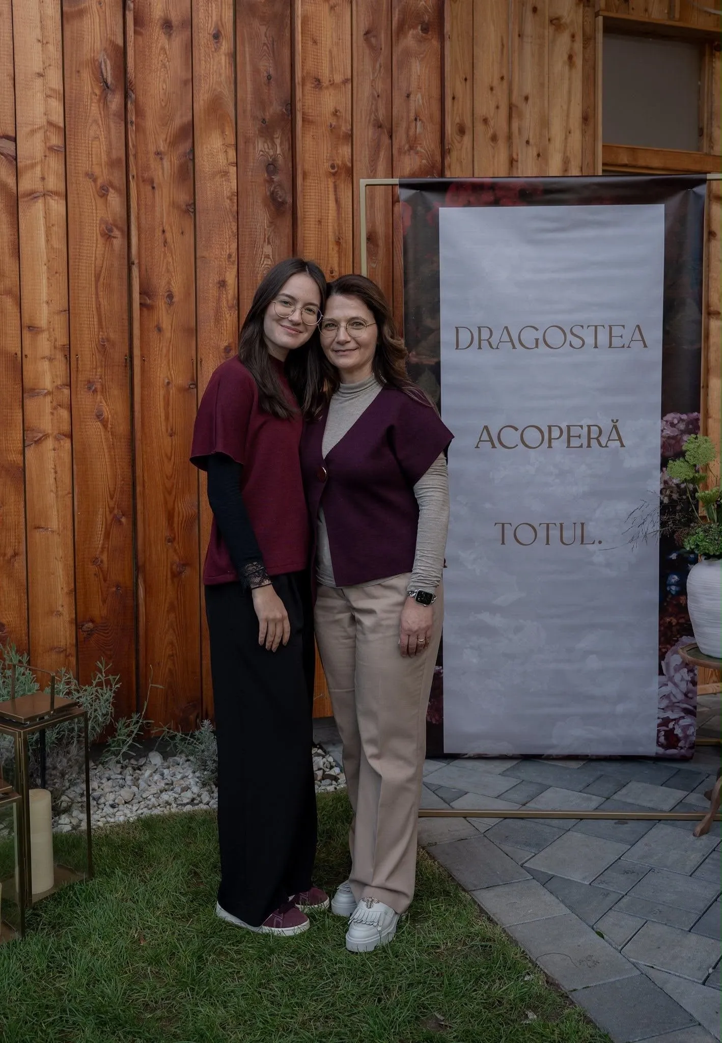 Two women in glasses and burgundy tops standing close together and smiling in front of a wooden wall, beside a banner that reads “DRAGOSTEA ACOPERĂ TOTUL.” (“Love covers all.”)