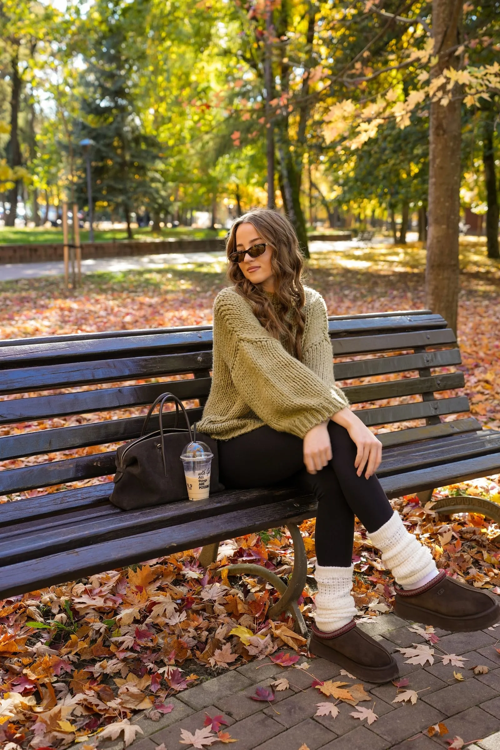 O tânără cu ochelari de soare și pulover verde-oliv tricotat, așezată pe o bancă de lemn într-un parc, printre frunze de toamnă