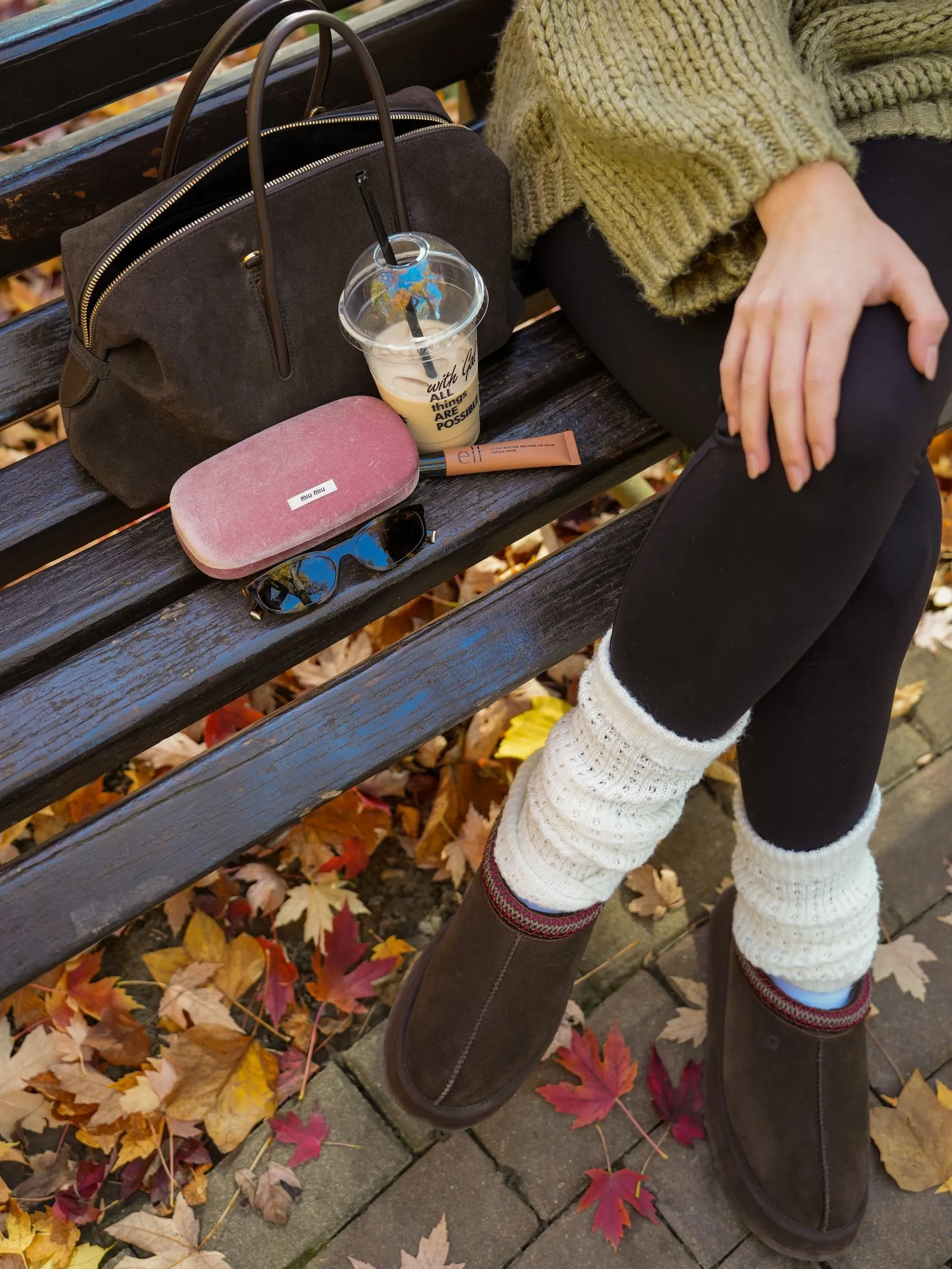 Detaliu pe o bancă din parc: o geantă din piele întoarsă, o cafea cu gheață, o cutie roz de ochelari și ochelari de soare, lângă jambiere tricotate și cizme maro, printre frunze de toamnă