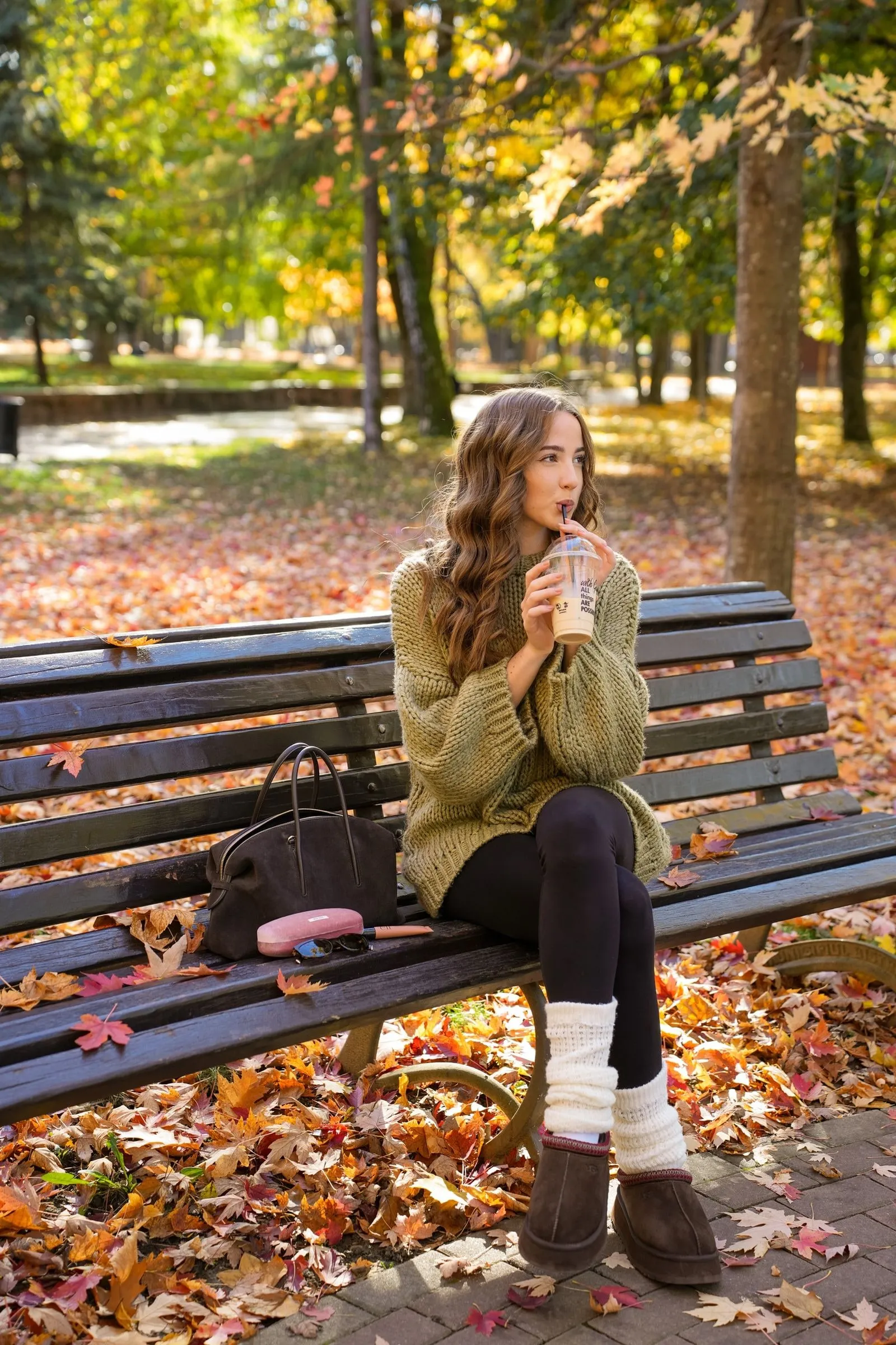 O tânără în pulover verde-oliv tricotat bea o cafea cu gheață pe o bancă din parc, înconjurată de frunze de toamnă