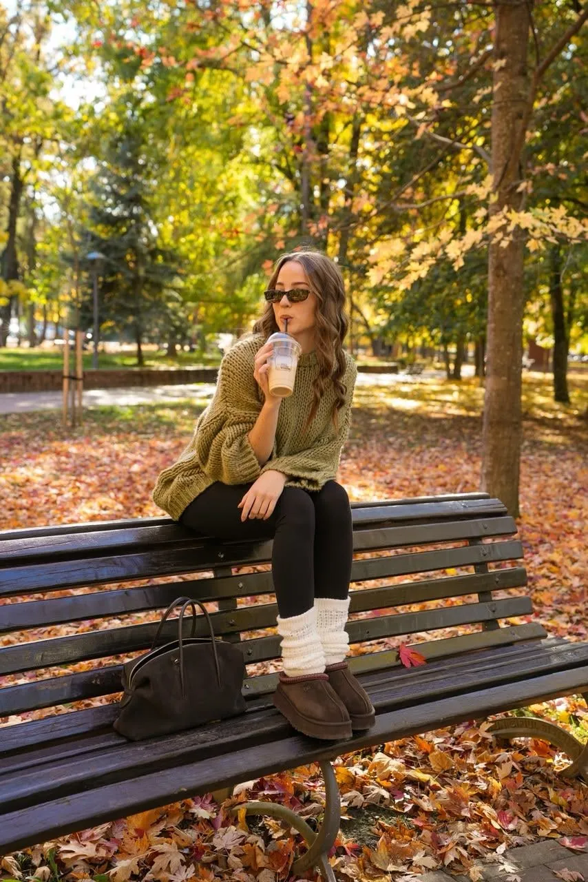 O tânără cu ochelari de soare și pulover verde-oliv tricotat, așezată pe spătarul unei bănci din parc, bea o cafea cu gheață, printre copaci și frunze de toamnă