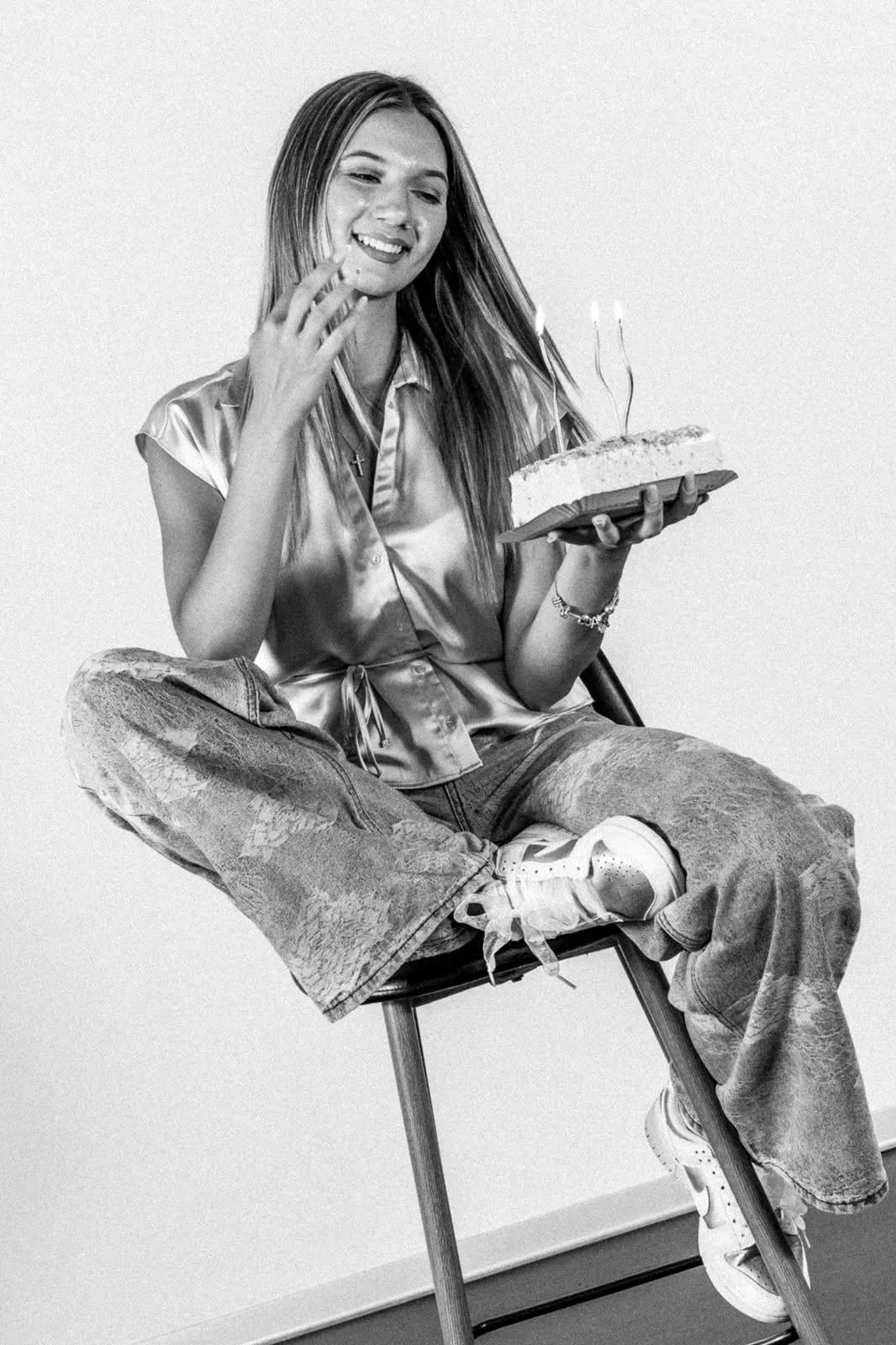 Black and white: a young woman laughing on a high stool, holding a cake with lit candles, against a plain light backdrop